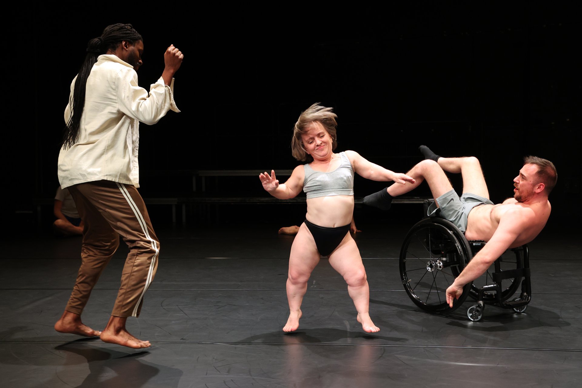 Drei Performer*innen mit unterschiedlicher Körperlichkeit. V.l.n.r.: ein Schwarzer Mensch im Sprung fotografiert. Eine kleine Person im Sprung, sie trägt Unterwäsche. Ein Mensch mit grauen Boxershorts, kopfüber im Rollstuhl sitzend.
