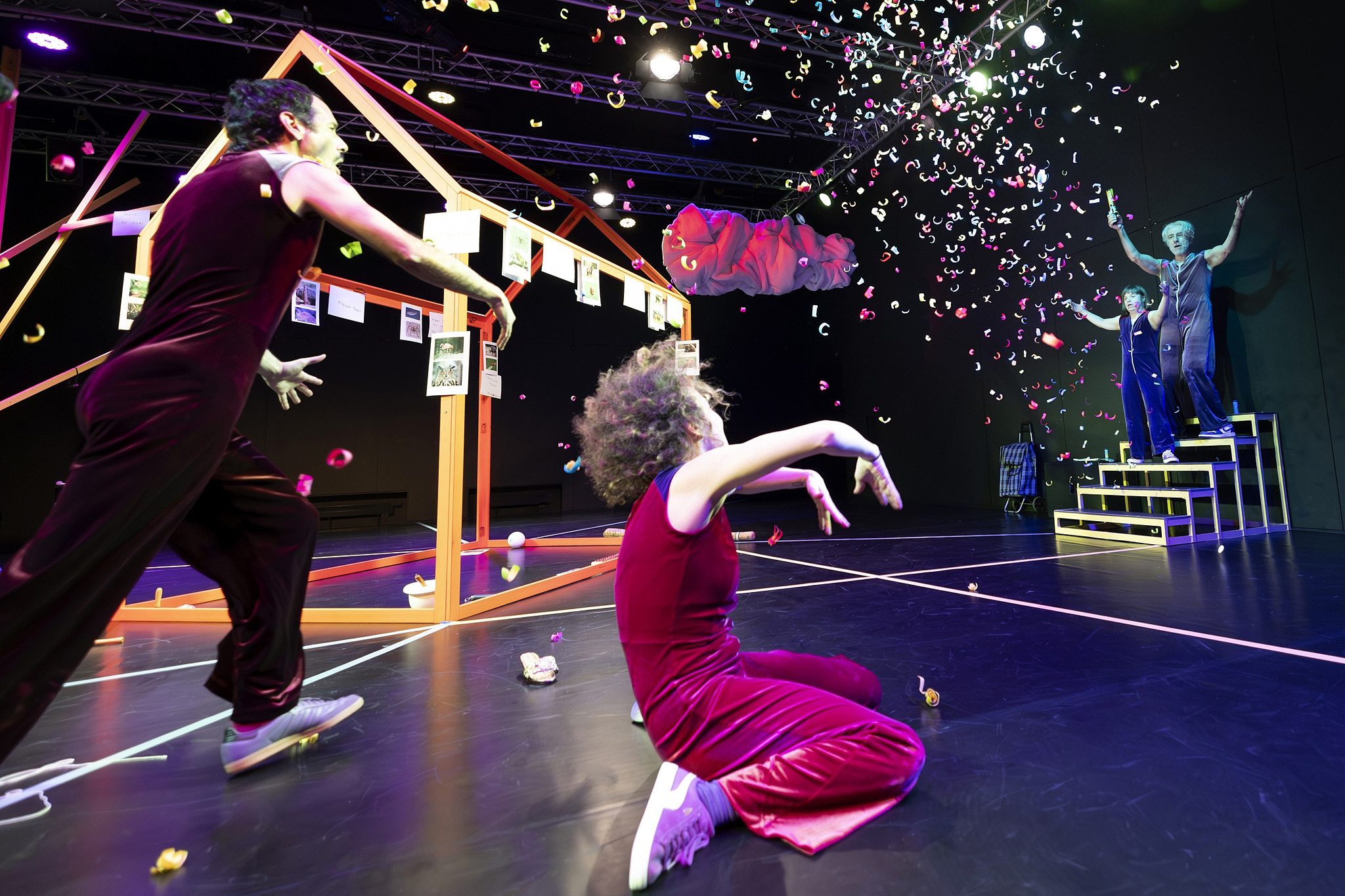 Im Vordergrund zwei Tänzer*innen, die zur Bühne gedreht sind. Auf der Bühne steht ein Holzgestell in Form eines Hauses. Rechts stehen auf einem Treppenpodest zwei Menschen, vor denen Konfetti fliegt.