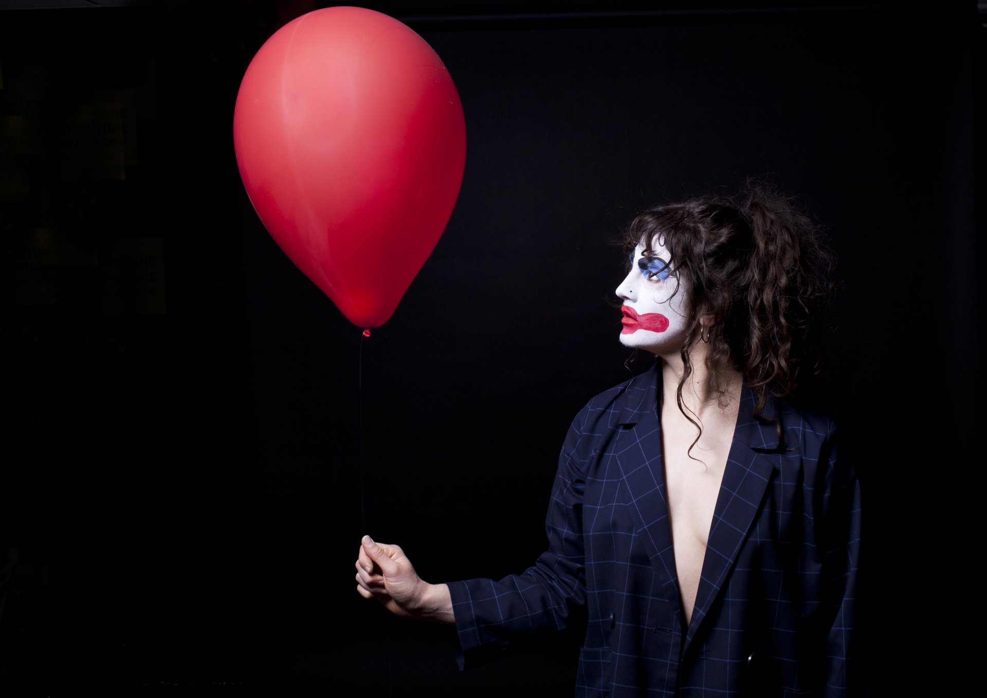 Eine Person hält einen roten Luftballon in der rechten Hand, der größer ist, als ihr Kopf. Sie trägt ein Nadelstreifen Sakko, hat die braunen Locken zum Zopf gebunden und trägt Clownsschminke (weißes Gesicht, übergroße rote Lippen und blauer Lidschatten).