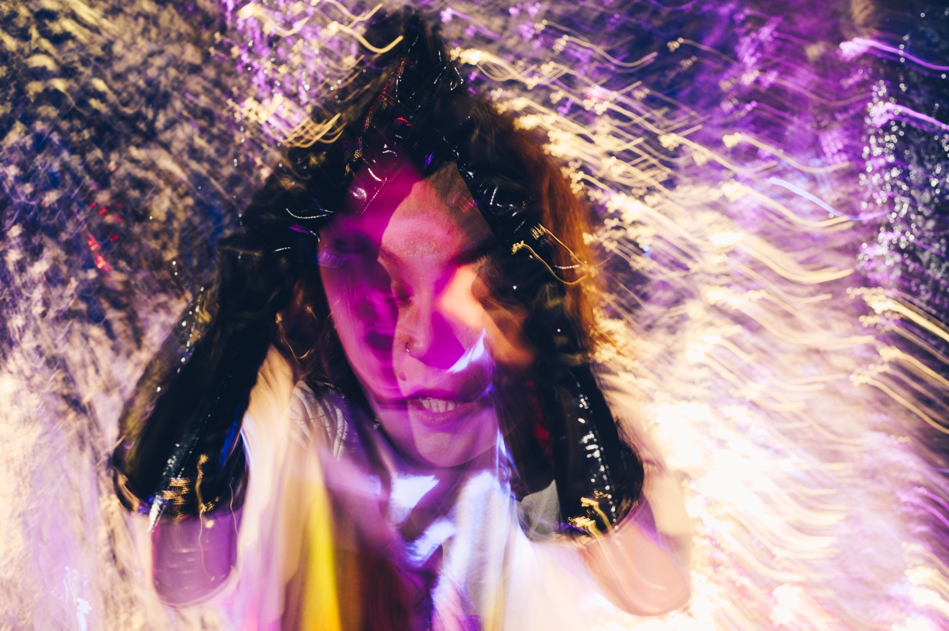 Eine Person mit langen Haaren ist inmitten von dynamischen Lichtstreifen in leuchtendem Lila und Gelb zu sehen. Das Bild ist stark verwischt und vermittelt durch diese Effekte ein starkes Gefühl von Bewegung. Die Person hält sich zwei schwarze Objekte an