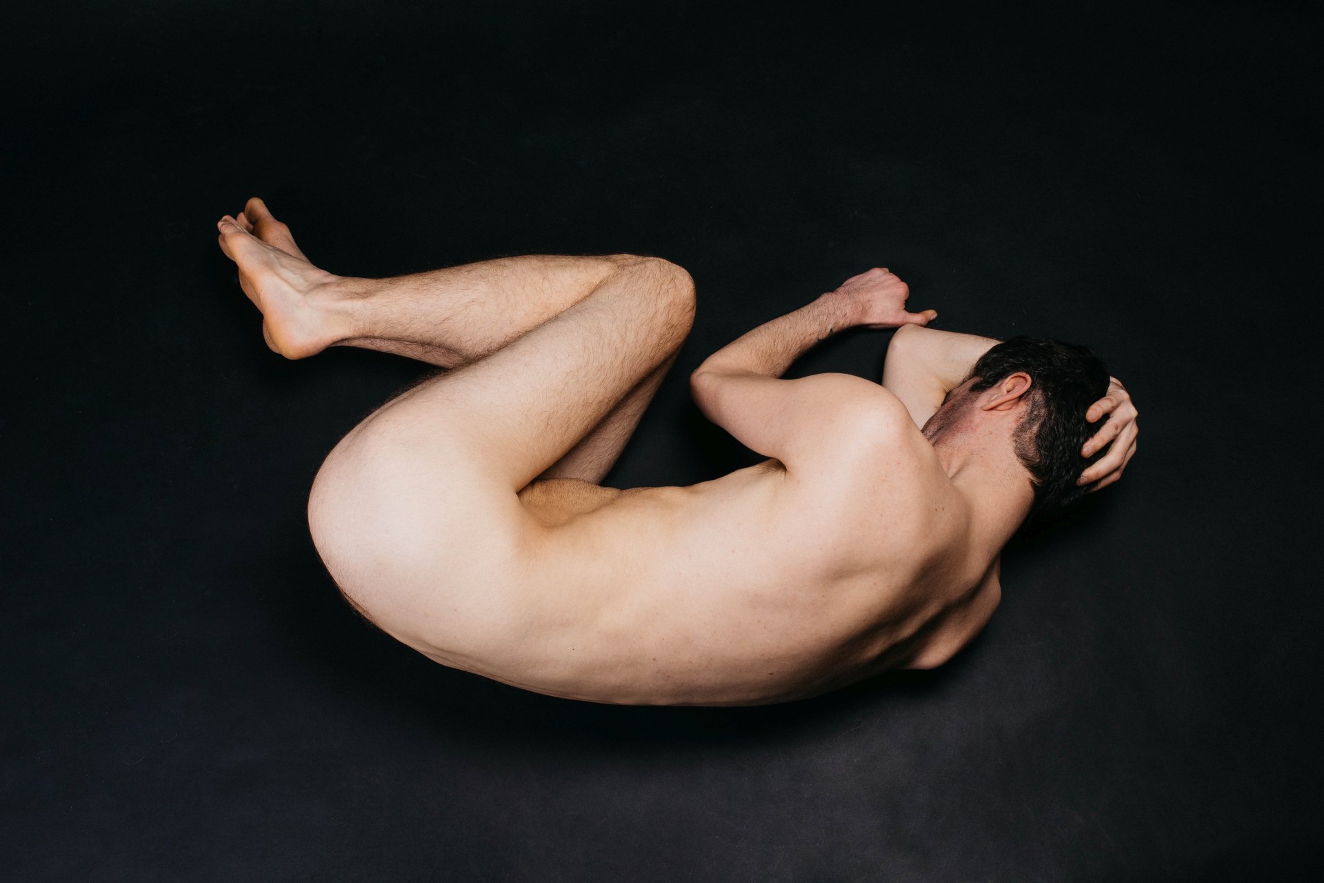 Eine nackte Person liegt seitlich zusammengerollt auf einem komplett schwarzen Untergrund. Das Gesicht ist in den Armen verborgen und die Beine sind eng an den Körper gezogen.