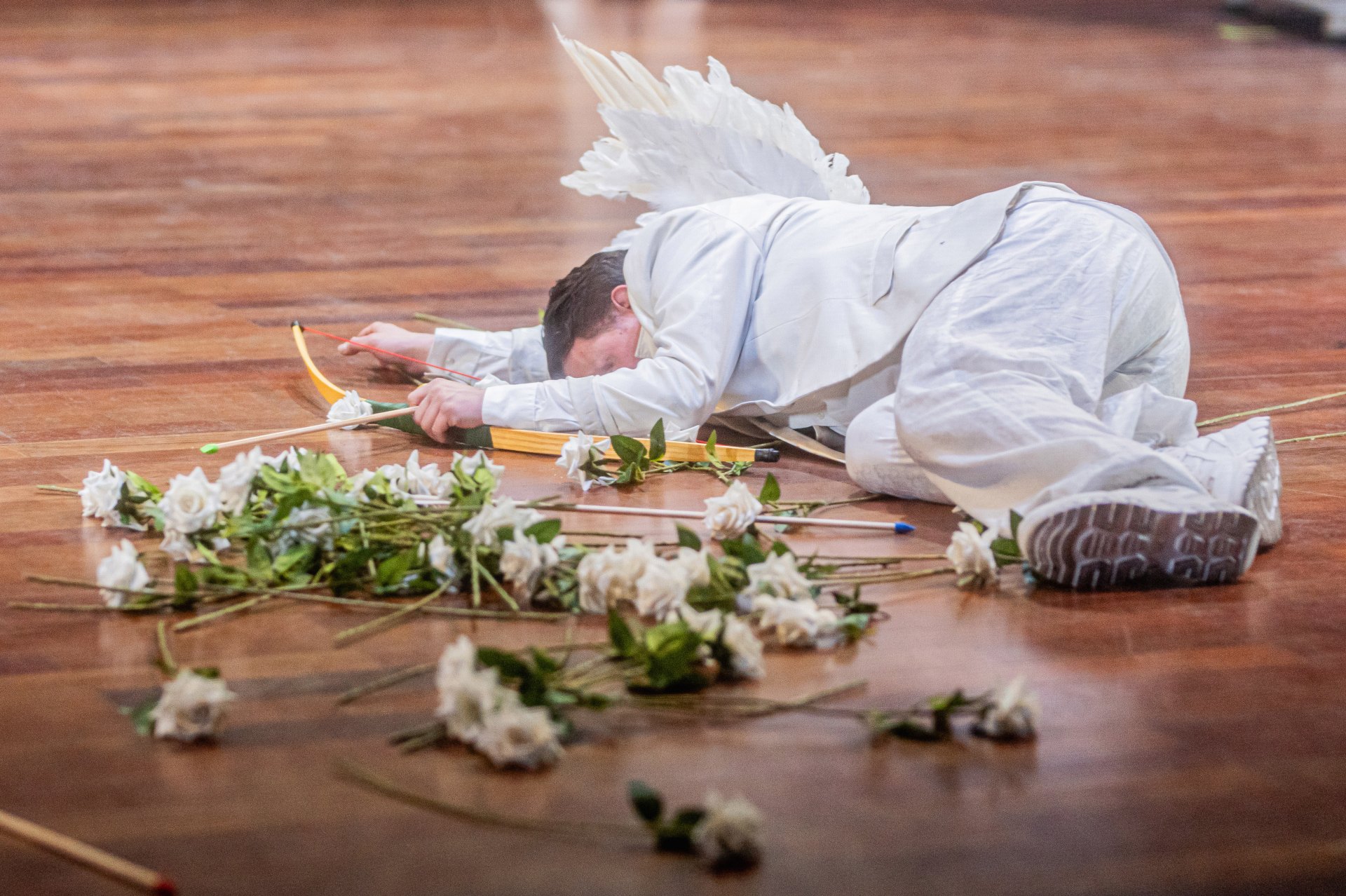 Eine Person in einem weißen Anzug und mit großen weißen Engelsflügeln liegt bäuchlings auf einem Holzboden. Sie hält einen Bogen in der Hand, während um sie herum viele weiße Rosen verstreut sind.