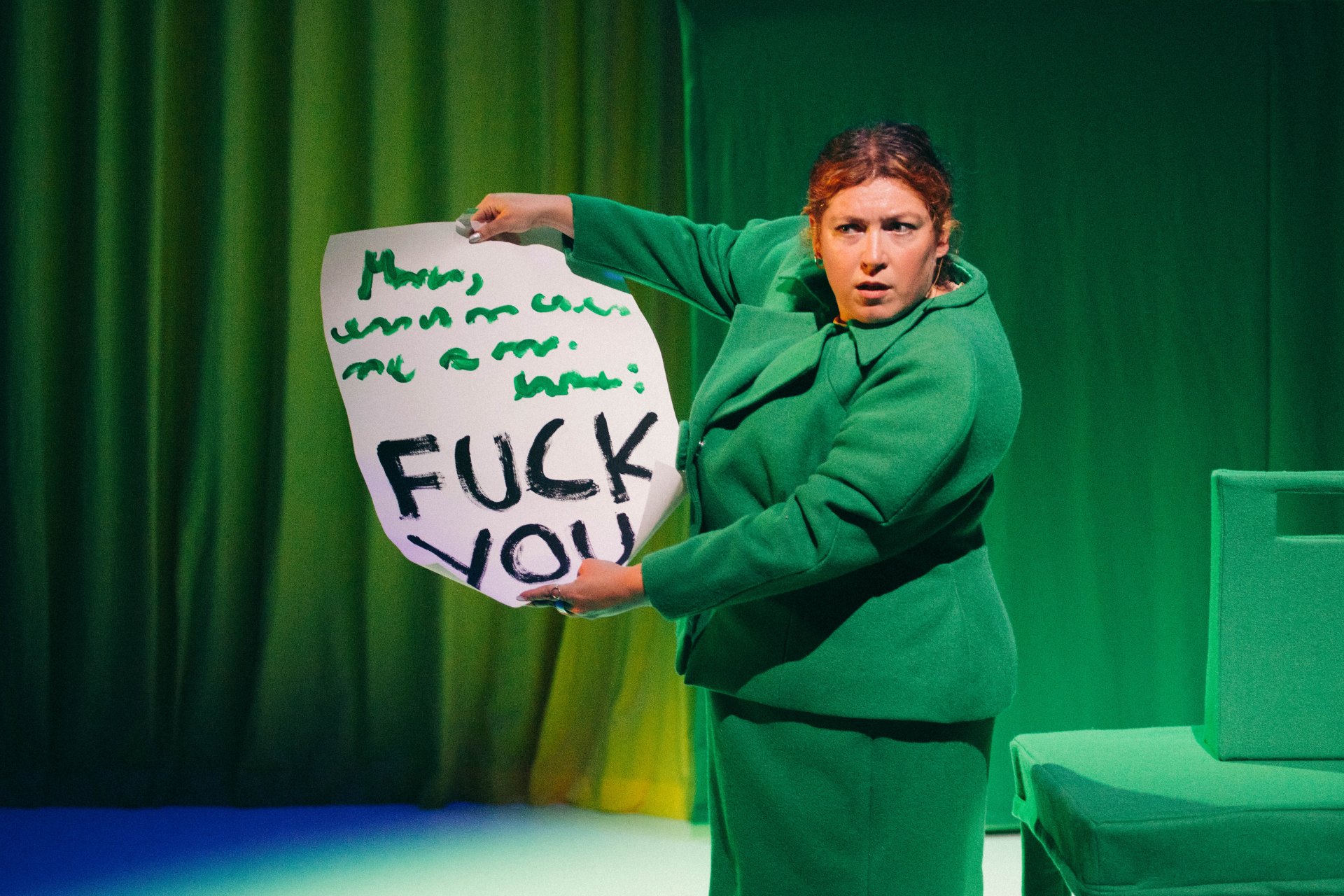 Eine Person in einem grünen Kostüm steht vor einem leuchtend grünen Vorhang. Sie hält ein großes Stück Papier hoch, auf dem deutlich in schwarzen Buchstaben "FUCK YOU" geschrieben steht. Der Gesichtsausdruck der Person ist dabei ernst.