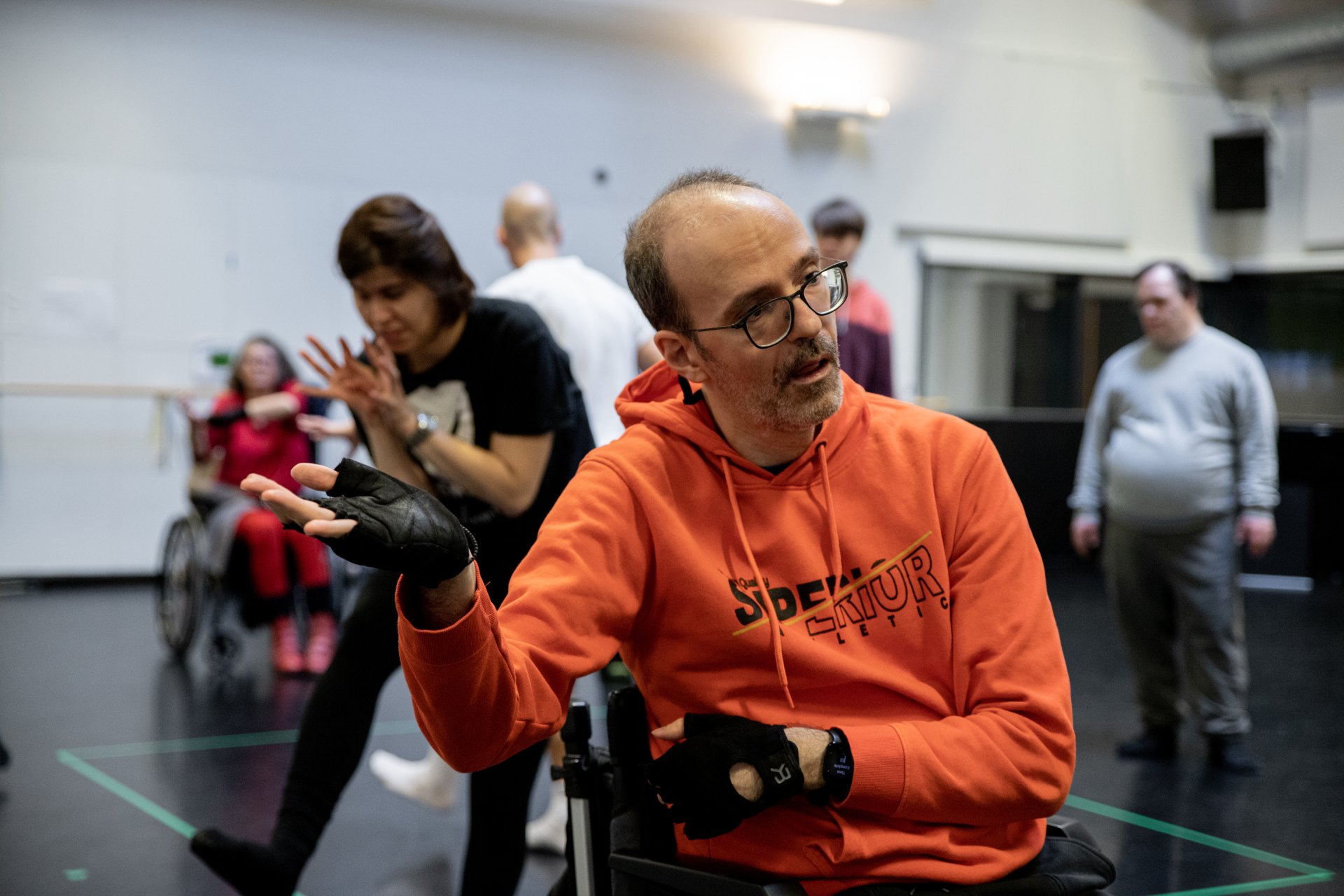 Eine Person im Rollstuhl mit orangefarbenem Pullover schaut nach rechts und hebt die Hand mit offener Handfläche nach oben. Im Hintergrund bewegen sich weitere Personen in einem Probenraum. Die Szene wirkt lebendig und partizipativ.